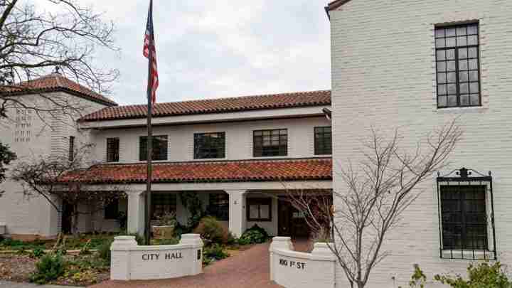 White brick City Hall with a red-tile roof, an American flag on a pole, brick walkway, and low front walls labeled 'CITY HALL' and '100 FT ST'
